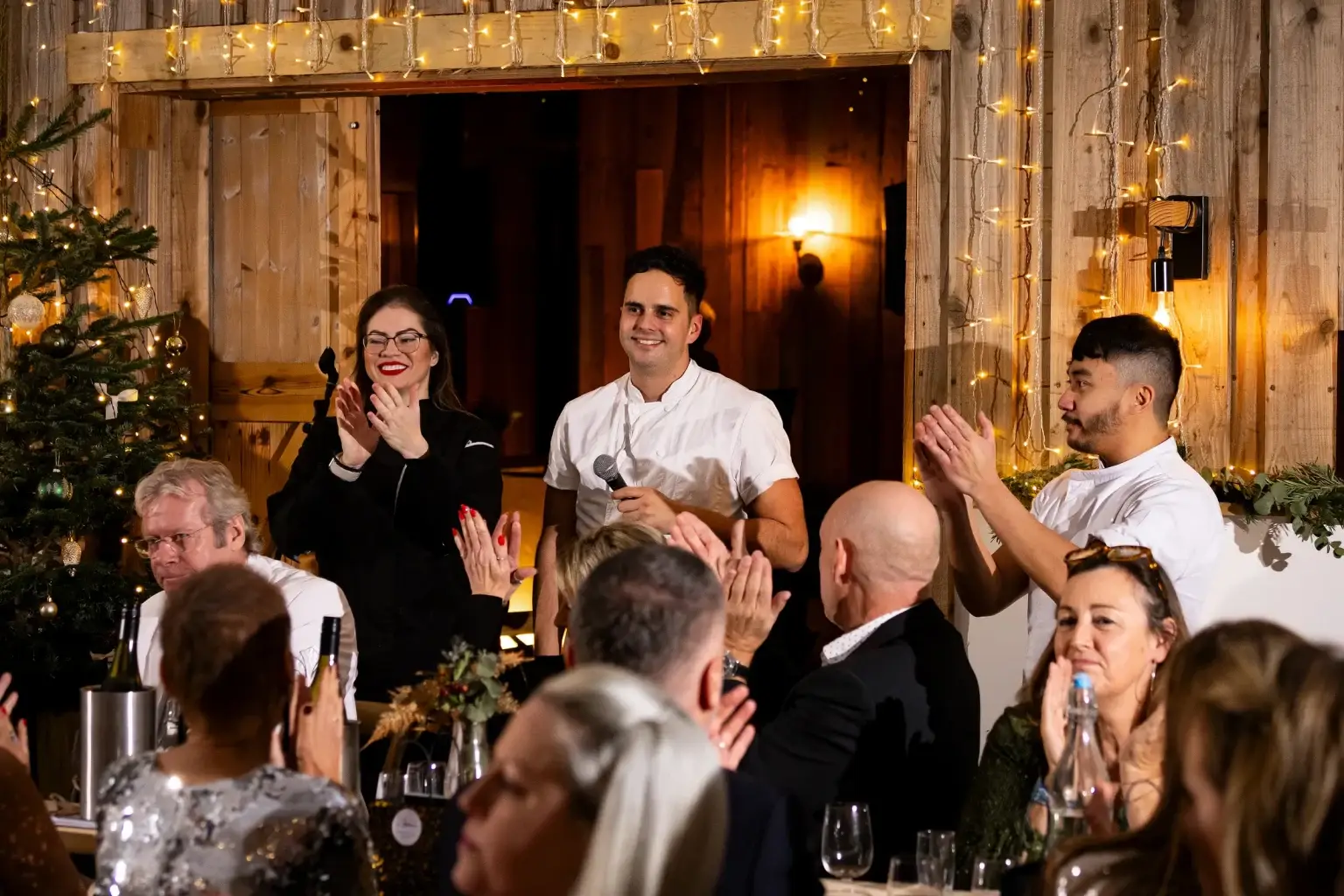 Nick Rapson holding a microphone beside the hospitality team as guests applaud around a dining table.