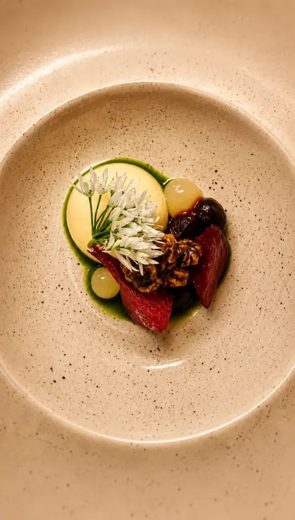 An overhead dish with dark red pieces, green sauce and white flowers in a warm ivory ceramic bowl.