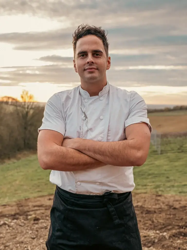 Chef Nick Rapson in a white chef jacket with folded arms outdoors at sunset.